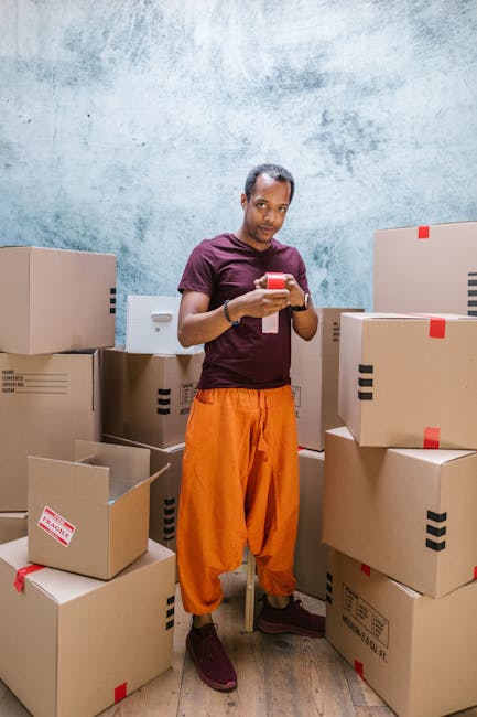A man and a woman are standing at the back of a white moving van, which is parked outside a residential property with a partly visible house and garden in the background. The man, dressed in a white t